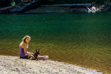 An adventurous athletic female hiker sitting on the shore of an alpine lake, looking at the camera smiling, in the Pacific Northwest with her French Bulldog.
