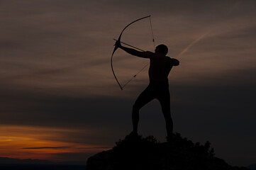 Silhouette of a handsome man with an ancient weapon bow and arrow on a background of sky and sunset.