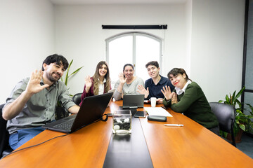 Group of employees sitting in office meeting room looking at camera and waving. Online meeting...