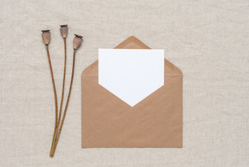 Blank white card mockup in brown envelope with poppy boxes, beige linen textile background. Top view, flat lay. Wedding invitation card.