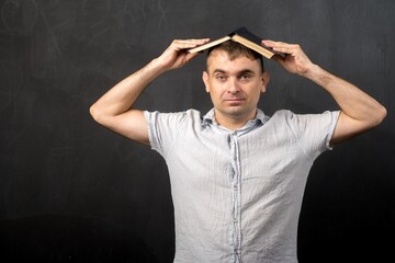 a man of European appearance stands against a black wall with a book on his head. tired teacher or...