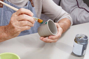 An elderly female hands paints a ceramic pot. The concept of creative leisure and development of the elderly. Close-up of a painter's hands. Selective focus
