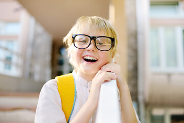 Smart little child with backpack on the stairs of school building. Quality education for children. Portrait of funny nerd schoolboy with big glasses. Vision problems. Back to school concept. - obrazy, fototapety, plakaty