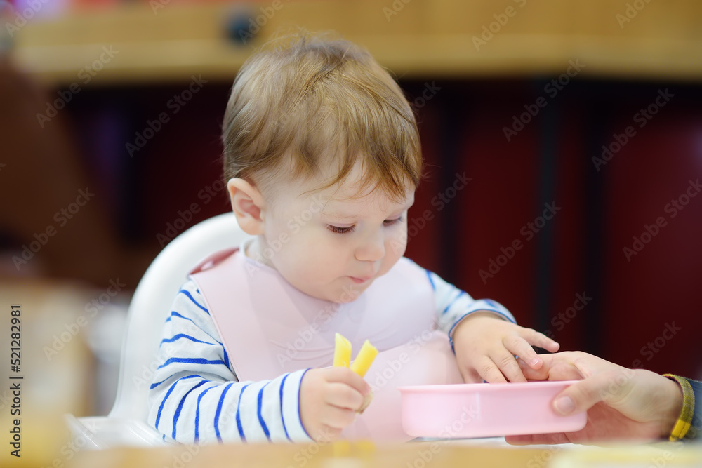 Wall mural Mother feeding cute baby boy healthy eating nutrition food in public place - airport or railway station. Childcare for small children during travel. Containers for baby food.