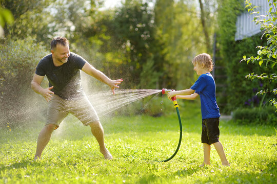 Funny little boy with his father playing with garden hose in sunny backyard. Preschooler child having fun with spray of water. Summer outdoors activity for family with kids.