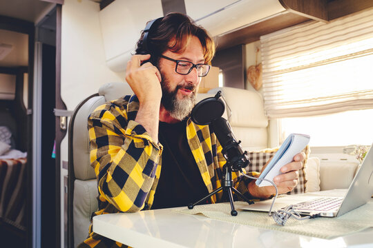 Portrait of young mature man with beard and glasses recording content online with a microphone in alternative van camper office house. Vanlife lifestyle people with modern job. Digital nomad life