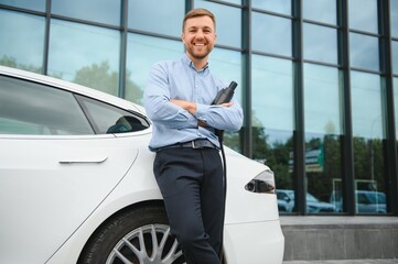 portrait of young handsome man in casual wear, standing at the charging station. Eco electric car concept.
