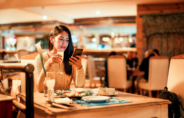 beautiful latin woman sitting at a table holding a cell phone and glass of drink in an elegant restaurant