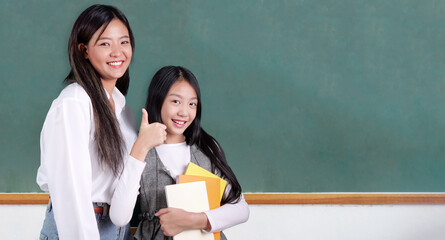 Happy teacher and schoolgirl with textbook on hand posing to camera with smile in the classroom,...