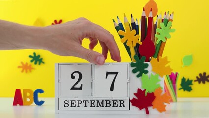 Wooden calendar with the date of September 27 on a table with a yellow background and a school decor, a woman's hand sets the date on the calendar. Concept background back to school, new school year