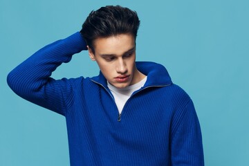 a young and slender man with thick black hair standing on a blue background in a blue zip-up sweater with a white T-shirt under it.Horizontal studio shot.