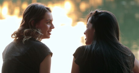 Friends talking by a lake. Girlfriends exchanging ideas and in conversation speaking to one another...
