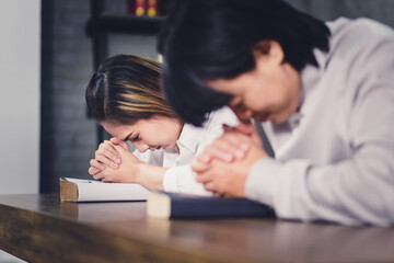 Young Christian couple wearing white shirt and praying at home. Catholic Asian man and woman sitting at table and praying  together with bible at church. 