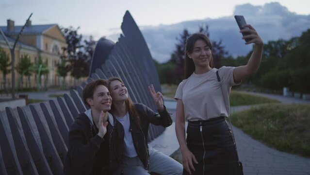 A group of friends is sitting in the park, the woman is taking pictures of everyone on the front camera, taking a selfie, friends are waving, grimacing, laughing. Summer evening.Slow motion 4k footage