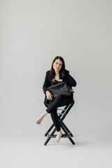 A girl in a black suit with a bag on a white background sits on a chair