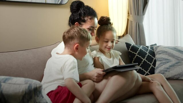 Happy family mom and cute little kids daughter and son laugh use smart tablet funny face mask app look at cell screen, mother with children watch cartoons make video call on mobile sit on sofa at home