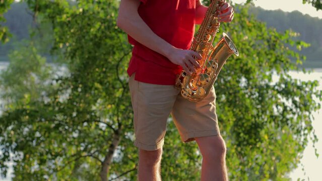 A young guy plays the saxophone near the river bank at sunset. Close-up.