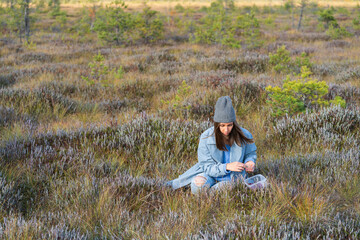 Naklejka na ściany i meble Young woman relaxes sitting on autumn swamp grass. Stylish girl in hipster hat collects ripe cranberries into small container. Female travels outside city for recreation and unity with nature