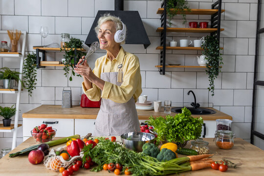 Senior attractive woman listening to music and singing in her headphones in the kitchen. Active lifestyle of mature people, enjoying life, having fun. Using whisk as microphone while cooking food