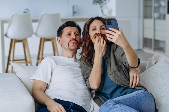 Cheerful in love couple taking a selfie on the phone while sitting on the couch together, making funny mustaches out of their hair and taking pictures