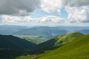 View of the green forest and grassy meadows of the hillside with the mountain village Kolochava in the valley on a cloudy summer day. Carpathian Mountains, Ukraine