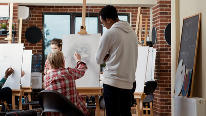 Man and woman giving advice to elder student in art class, drawing vase model on canvas to develop...