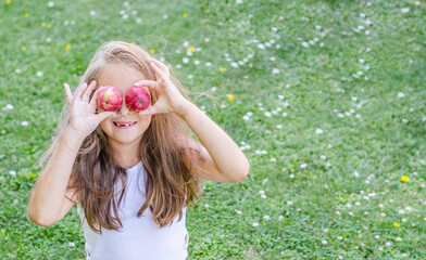 A little girl holds apples in her hands while sitting on the grass and covers her eyes.
