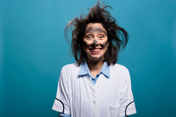 Portrait of crazy chemist with dirty face and messy hair grinning dreadful at camera while on blue background. Mad scientist with dreadful smile and stupid look after chemical laboratory explosion. - obrazy, fototapety, plakaty