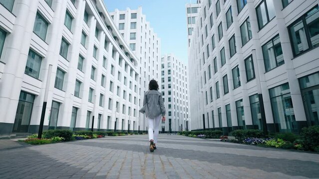 young woman is walking in modern district of big city, full-length moving shot, rear view