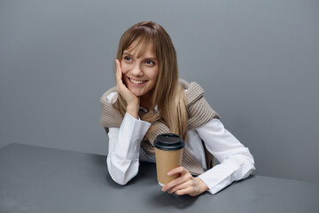 Smiling dreaming young blonde student lady freelancer in warm sweater with takeaway coffee recline on hand looks up in gray modern home office. Coffee break Lover Concept. Copy space. Cool offer