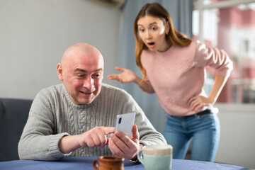 Man sitting at table at home and using smartphone. His jealous wife standing behind.