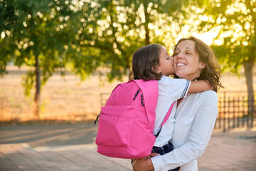 Fototapeta premium smiling long-haired brown-haired mother in a white shirt with her daughter in her arms with a pink backpack giving her a kiss