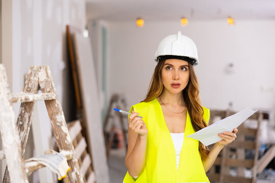 Young beautiful woman in uniform, standing on a construction site indoors during repair work, makes important notes on paper