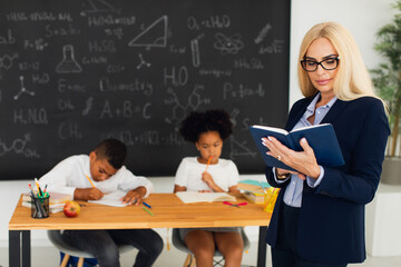 Portrait of female teacher standing on the background of a classroom and school children.