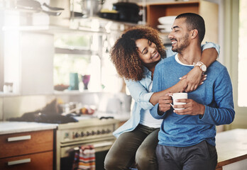 Romantic loving couple, smiling and enjoying a cup of coffee in the morning together at home in their kitchen. Carefree wife hugging and bonding with husband while relaxing looking in love