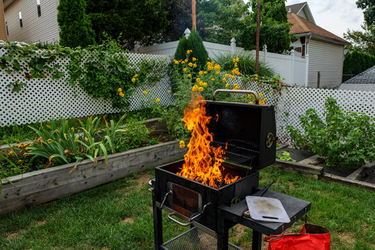 Brightly burning in metal box logs firewood for barbecue outdoor on sunny day. Orange high flame and white smoke on blurred blue sky and green grass background. Camping, safety and tourism concept