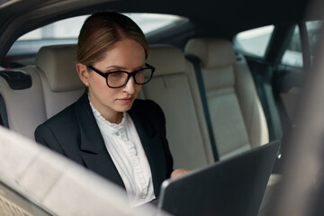 Serious Concentrated Business Woman 60-65 Years Using Laptop in Car. Female in Glasses Holding Computer PC in Lap and Type on the Keyboard