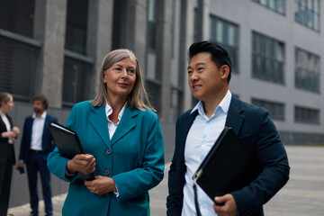 Man and Woman Colleagues Walking Discussing Documents. Colleagues in Elegant Suits Going on Street with Clipboard. Urban Background 