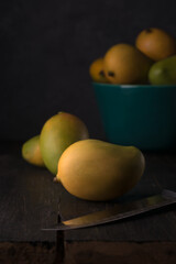 ripe mangoes, tropical fruit on table top, with bowl of fruits and knife, dark background with soft-focus, copy space