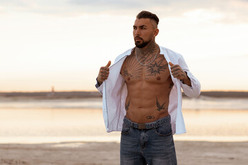 Muscular athletic sexy man in white shirt with bare-chested rests on beach. Portrait of tanned fit...