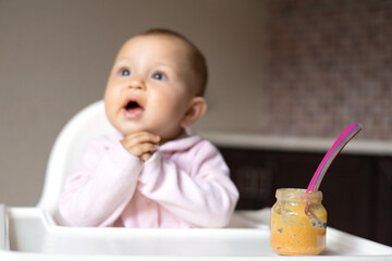 Baby food. A baby in a high chair eats vegetable puree from a spoon. Mom feeds the baby from a jar. Kitchen. Lifestyle.