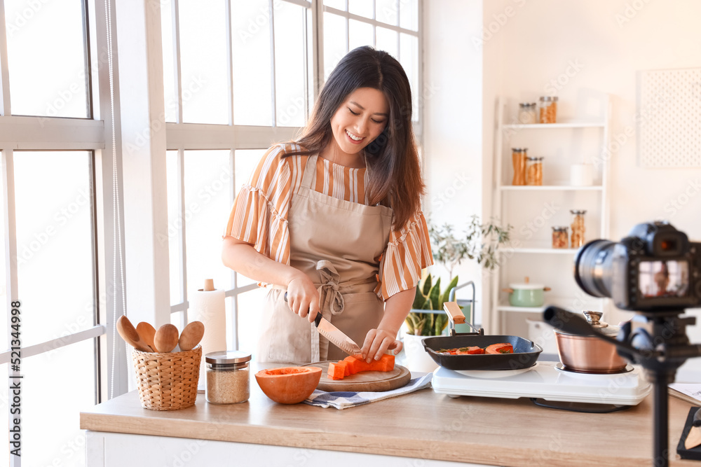 Sticker Young Asian woman cutting pumpkin while recording video class in kitchen