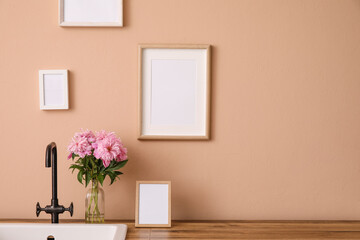 Fototapeta na wymiar Vase with peony flowers and modern sink on kitchen counter near color wall with blank photo frames