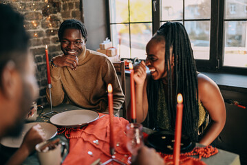 Best friends celebrating new year. Young people with candles, sitting at dining table. Diverse students during christmas party at home, smiling and laughing.