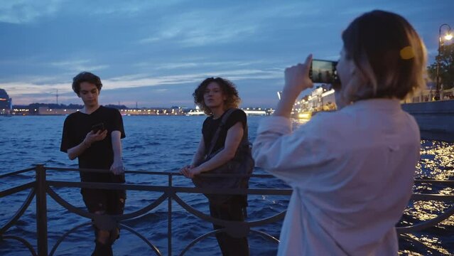 A group of friends stand on a pier near the river and take pictures. Summer night. Slow motion 4k footage