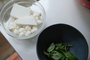 Fototapeta na wymiar Top view of fresh ingredients: mozzarella and salted feta cheese in a glass transparent bowl, aromatic basil leaves on navy ceramic bowl on white kitchen table. Copy ad space. Food, cuisine, culinary