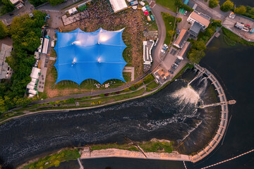 Concert of beer festival area with big blue tent and service areas by a river. Big crowd enjoy all amenities. Aerial top down view. Music event or circus in town. Entertainment industry and business.