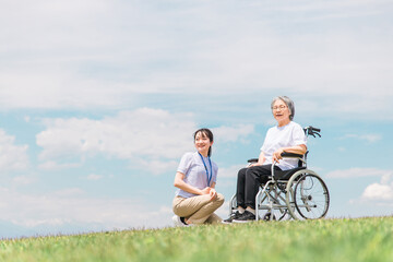 公園を散歩する介護士と車椅子の高齢者女性（リハビリ）
