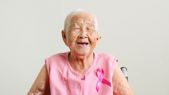 Portrait of senior Asian woman in pink shirt with pink ribbon smiling on gray background for October breast cancer awareness month