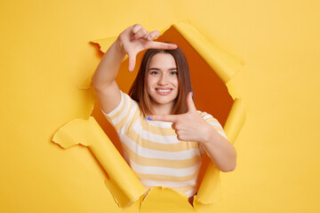 Indoor shot of smiling woman wearing striped t shirt looking through breakthrough of yellow background, gesturing photo, zooming focusing to camera, observing with happy expression